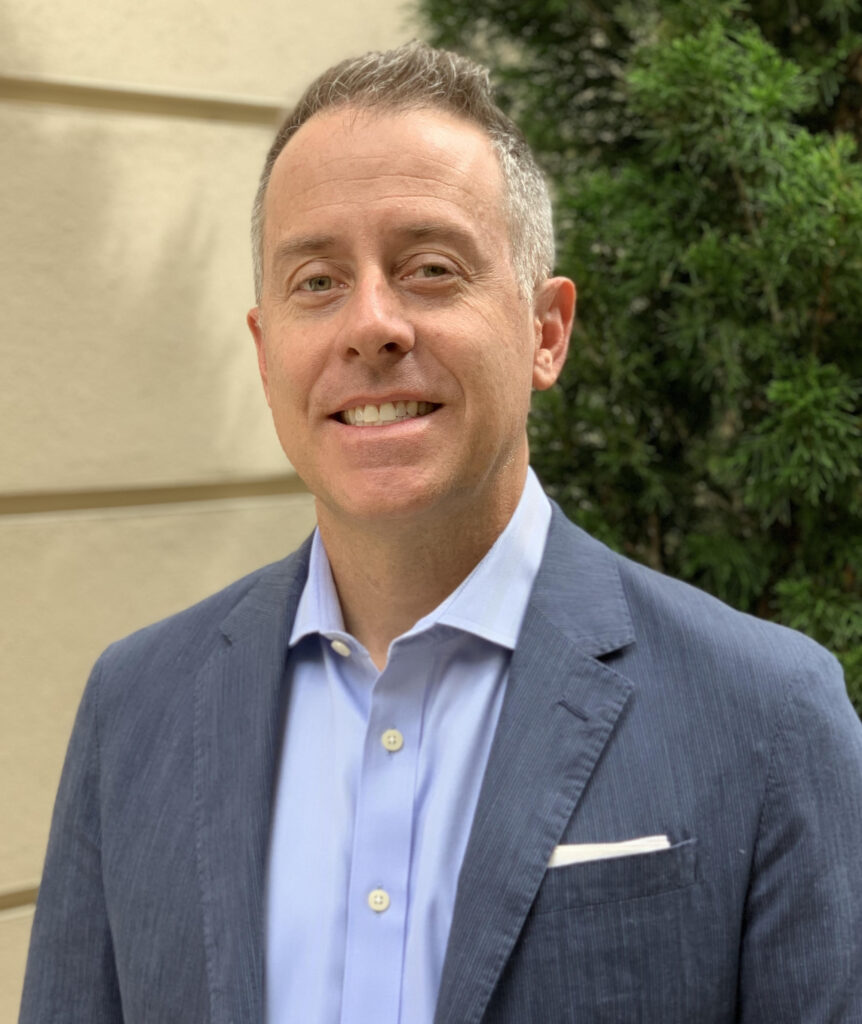 Smiling man in a blue blazer and light blue shirt standing outdoors with greenery in the background.