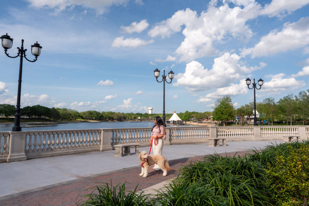 Woman walking a small tan poodle on a riverside promenade with ornate lampposts and a stone railing.