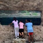 Three kids stand on a small rock platform, peering through a rectangular opening in a rocky wall toward a water-filled cave beyond.