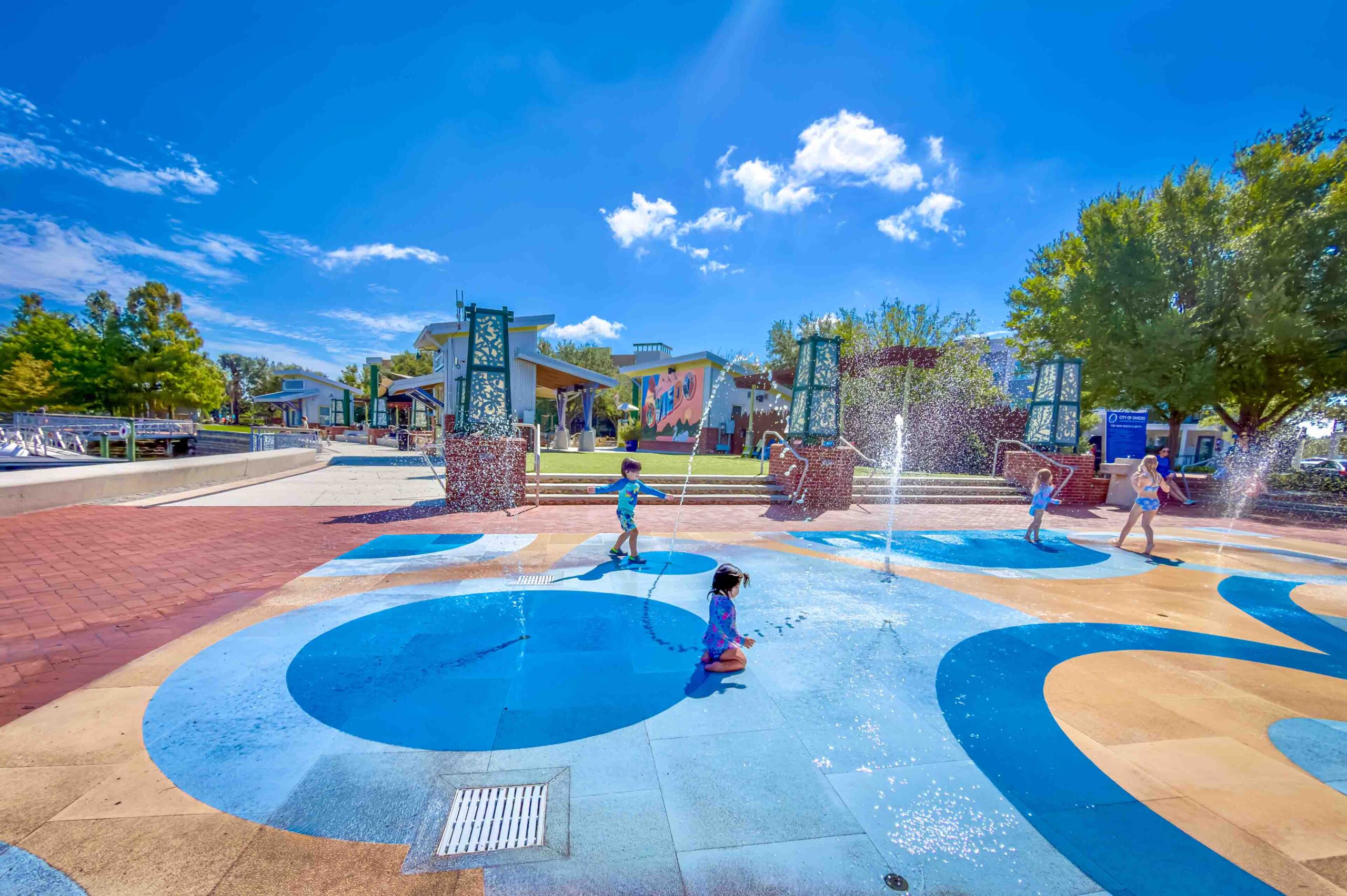 Children playing in a splash pad with water jets and blue circular tiles on colorful pavement in a sunny park.
